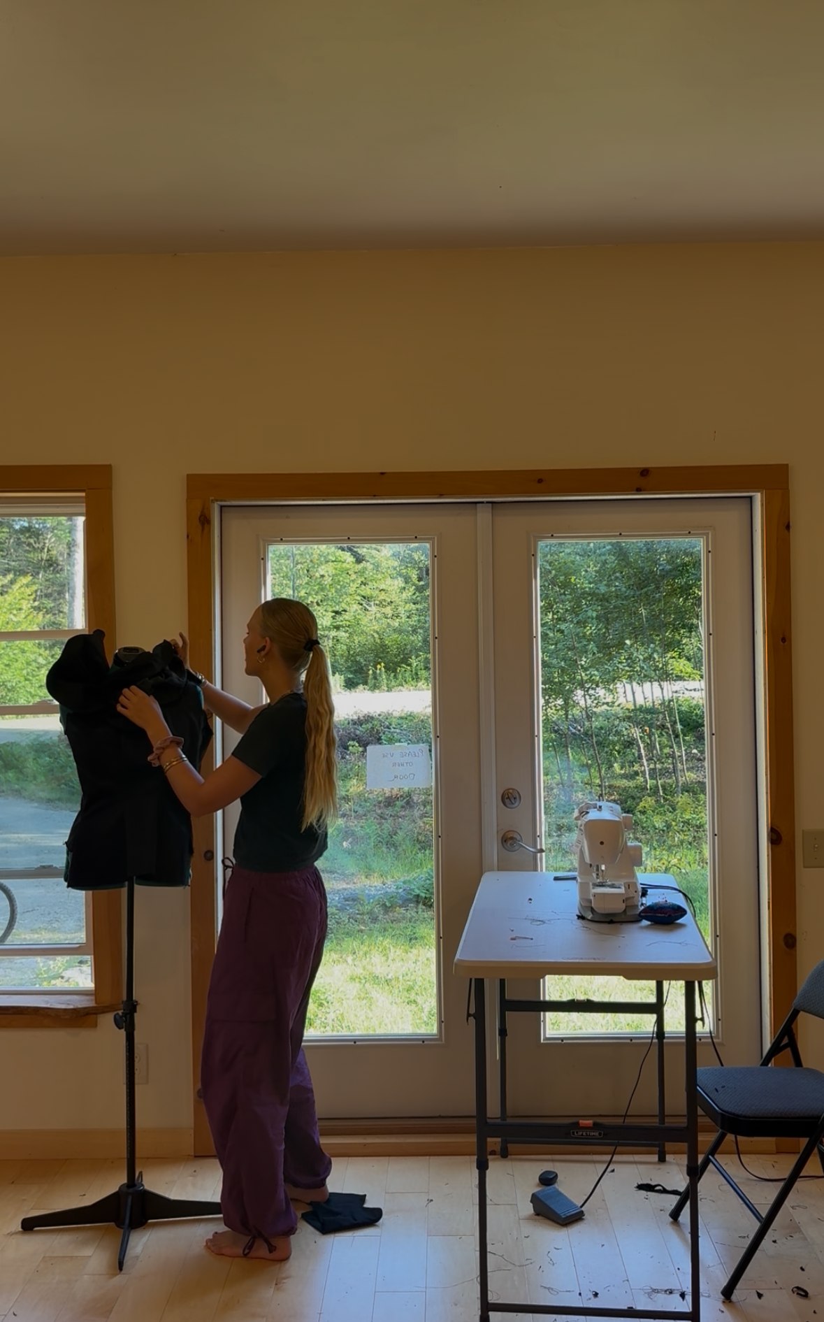 Seriena adjusting a black garment on a dress form in her sunlit studio, sewing machine in the background