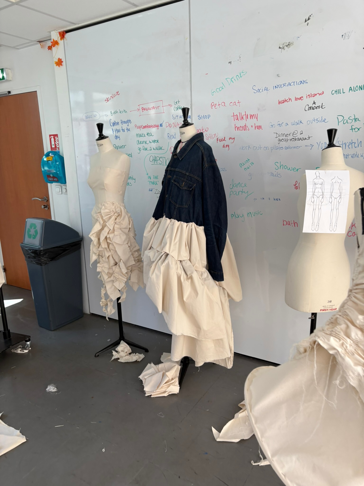 Design studio with dress forms wearing muslin garments in progress and an upcycled denim jacket, sketches and mood notes on the wall