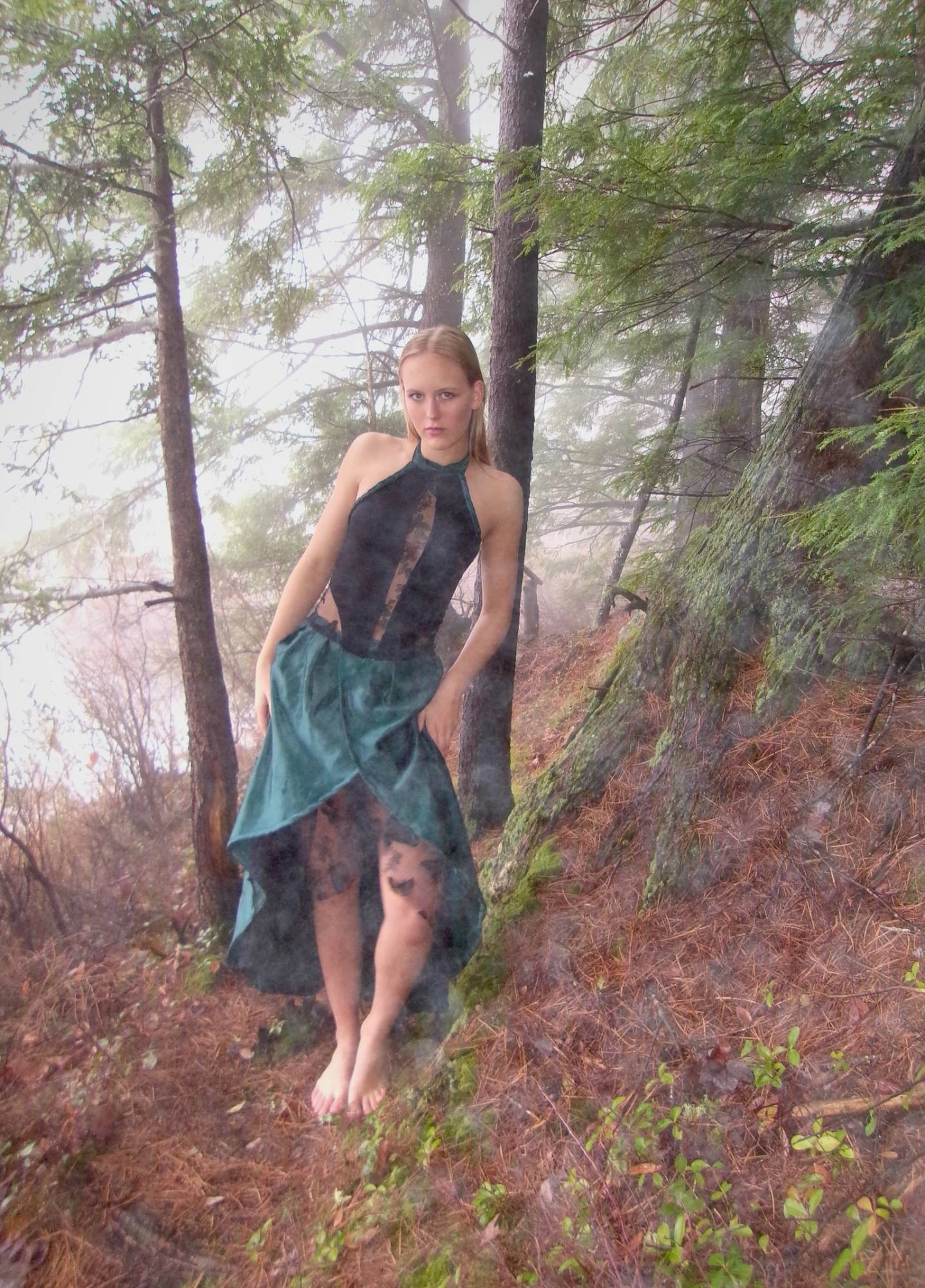 Model in a black bodysuit with a sheer teal wrap skirt, standing in a foggy forest