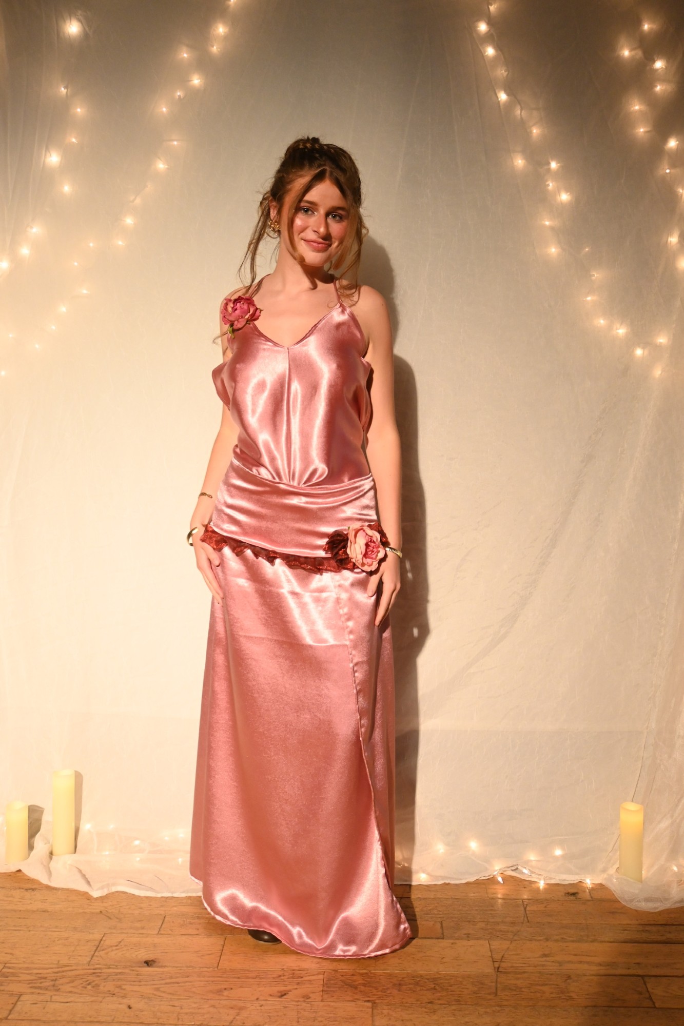 Model wearing a pink satin cami and skirt set with fabric rose details, fairy-lit backdrop