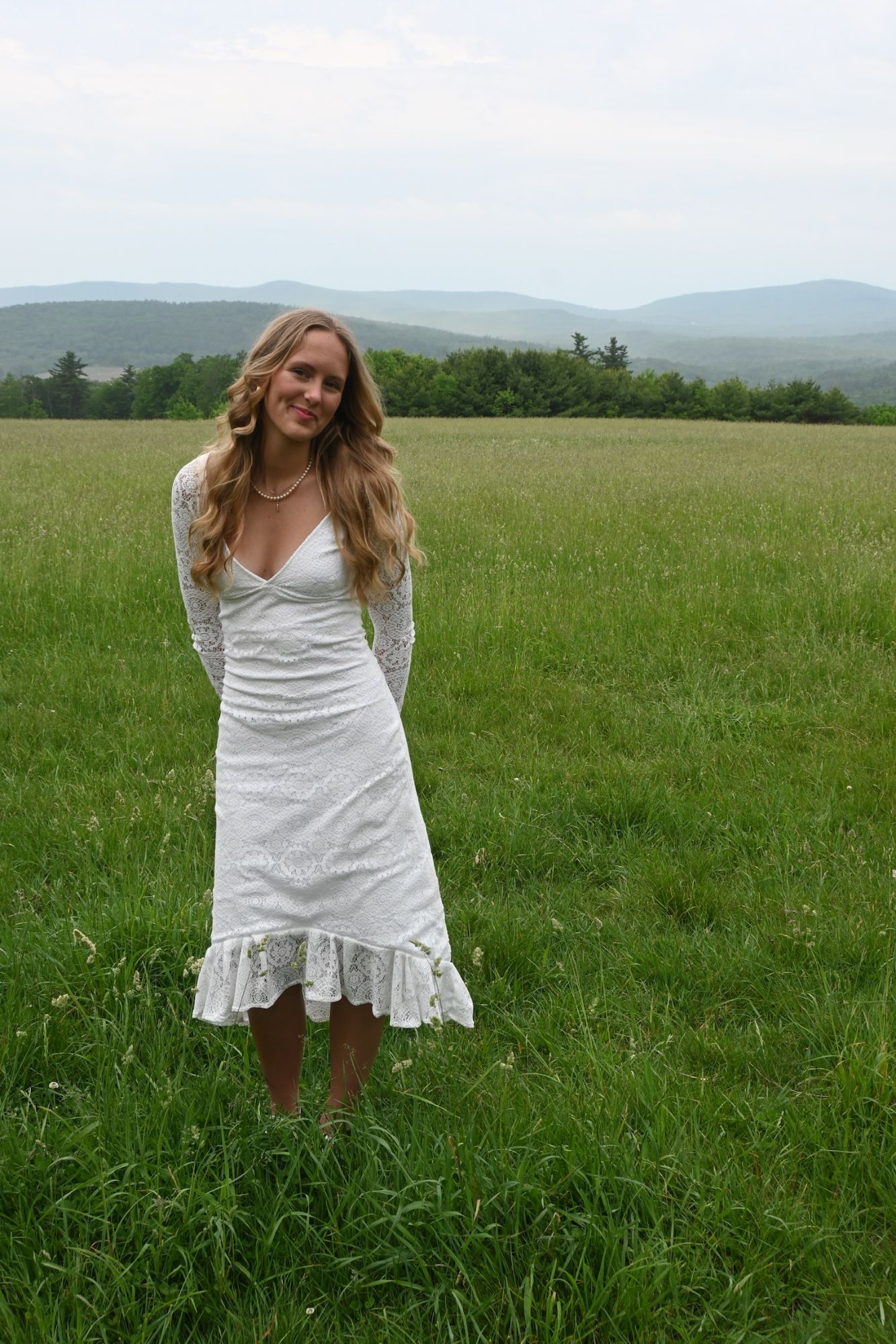 Model in a long-sleeve white lace dress standing in a green field with mountains behind